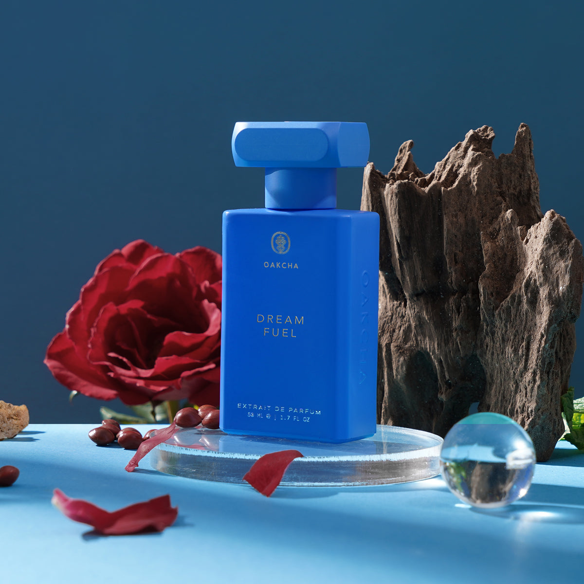 Front view of Dream Fuel blue perfume bottle with T-shaped cap on glass pedestal, red rose petals and driftwood on blue background