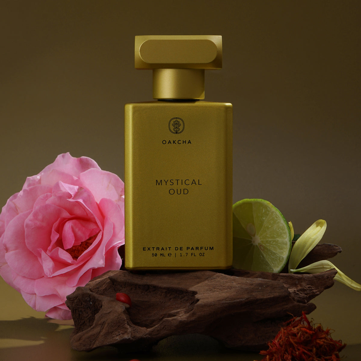 Front view of Mystical Oud perfume bottle showcasing gold rectangular bottle with logo beside a pink rose and agarwood on a brown background