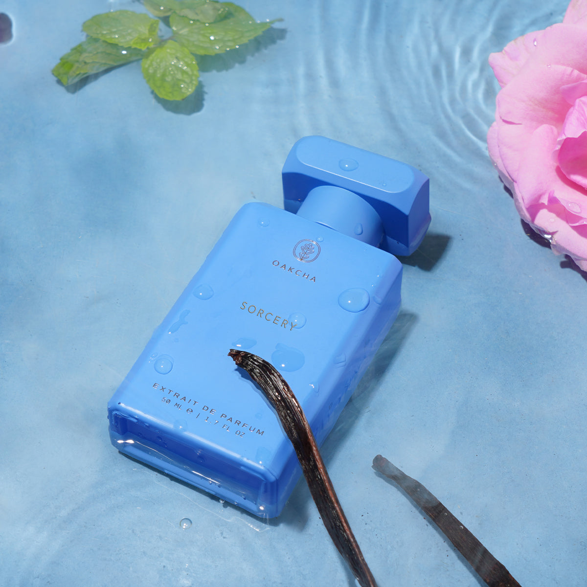 Three-quarter view of Sorcery bottle showcasing bright blue rectangular bottle with water droplets on a rippled blue surface and a pink rose nearby