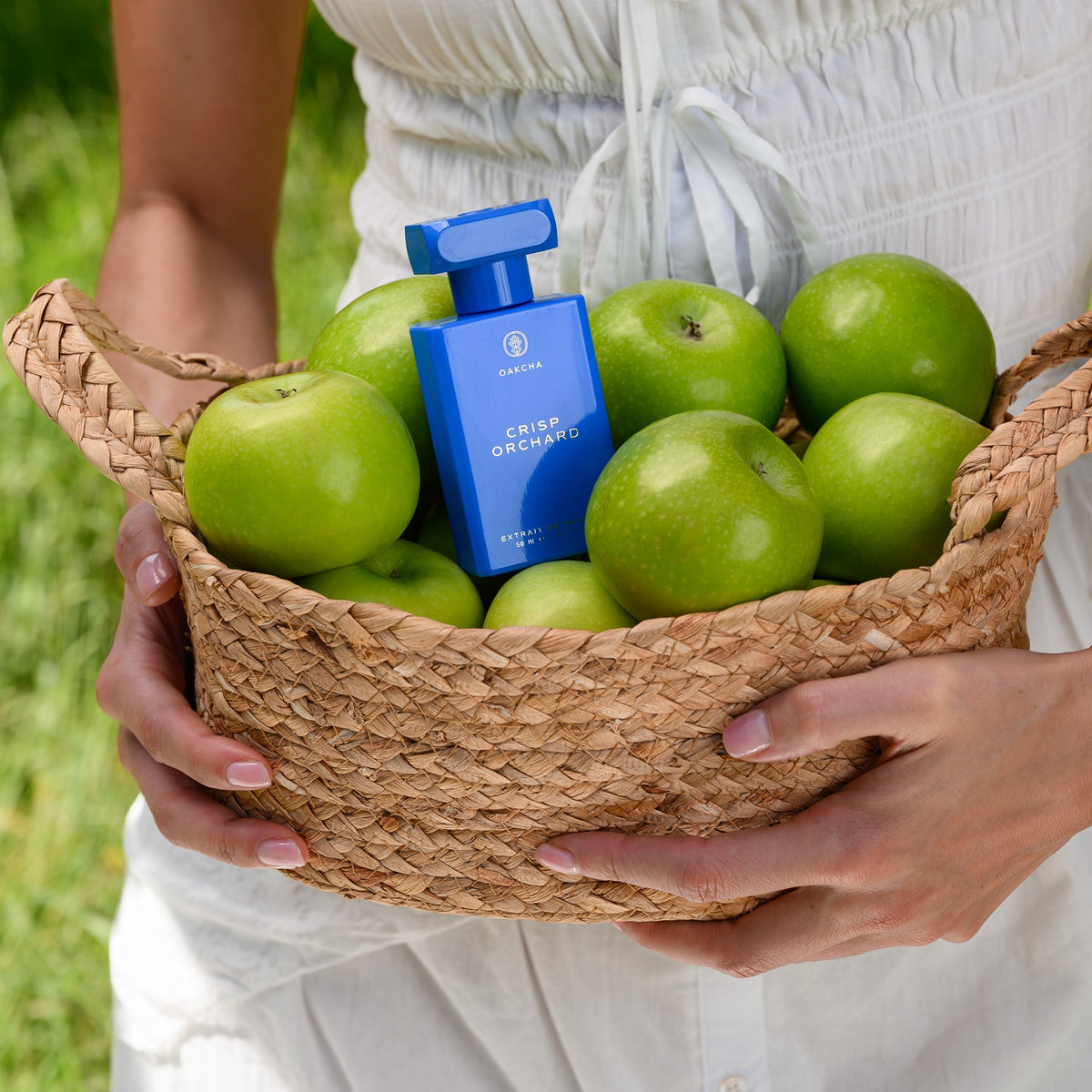 Blue Crisp Orchard Extrait de Parfum bottle in a woven basket of green apples held by a model in a white dress
