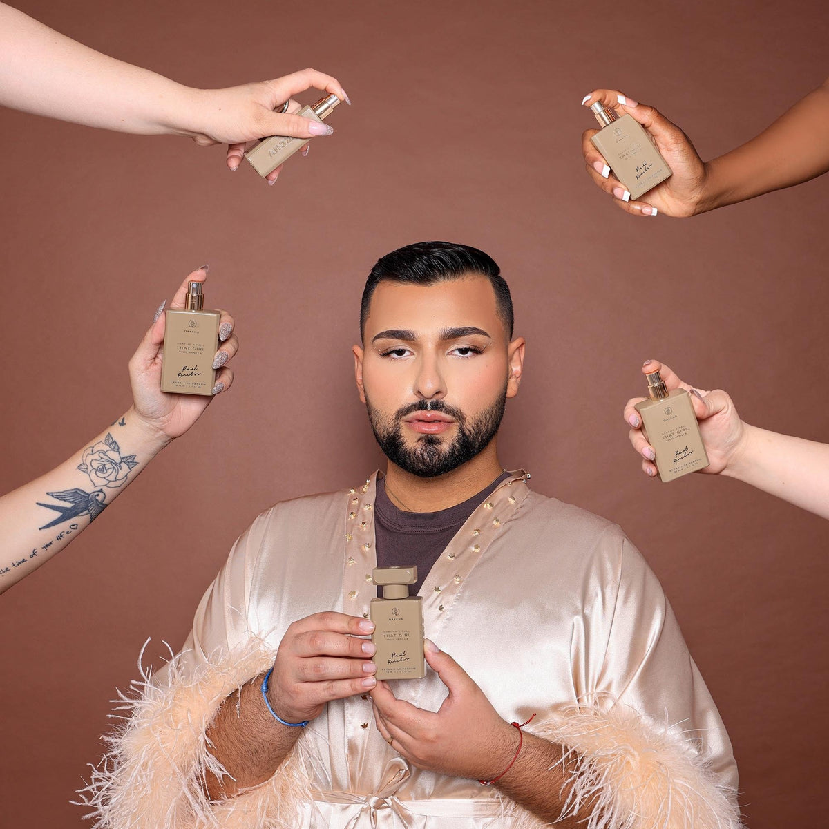 Man holding That Girl Viral Vanilla perfume with additional bottles around him, wearing a satin robe with feather cuffs