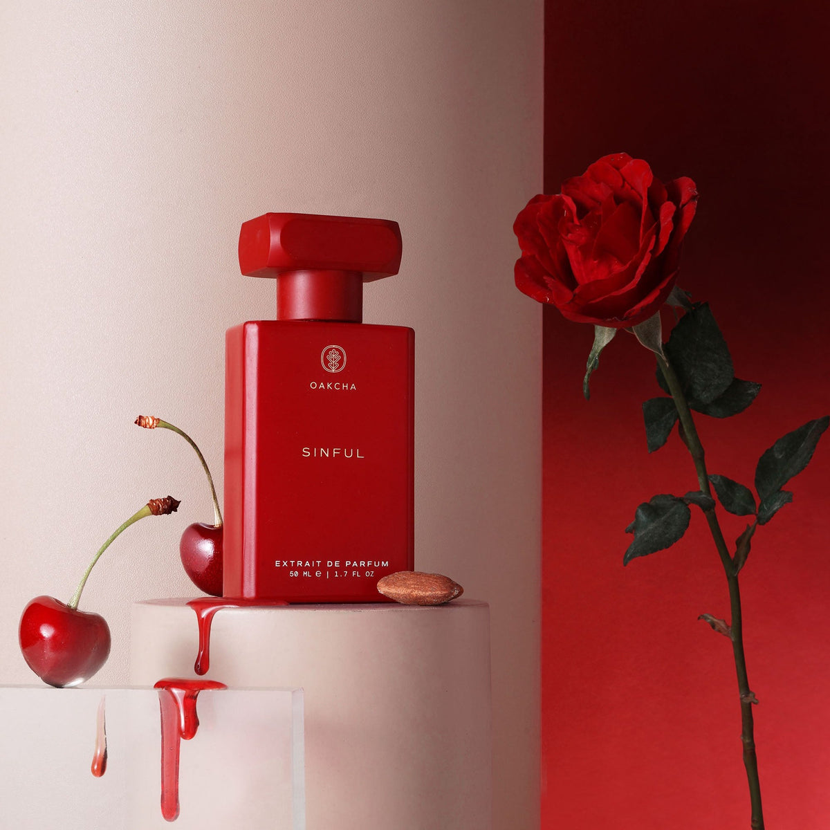 Front view of Sinful Extrait de Parfum showcasing its red bottle with cherries, a rose, and almond on sculptural pedestals